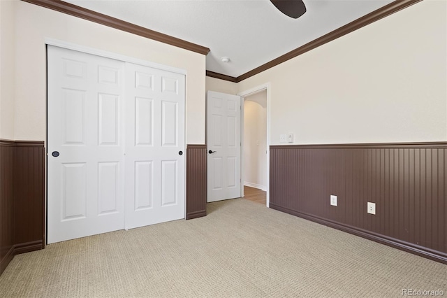 unfurnished bedroom featuring wainscoting, ornamental molding, light colored carpet, a closet, and a ceiling fan