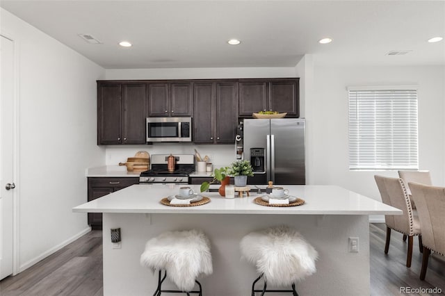 kitchen with dark brown cabinetry, wood finished floors, a kitchen breakfast bar, light countertops, and appliances with stainless steel finishes