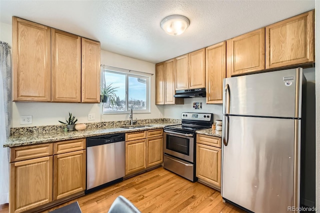 kitchen featuring light wood finished floors, appliances with stainless steel finishes, light stone counters, under cabinet range hood, and a sink