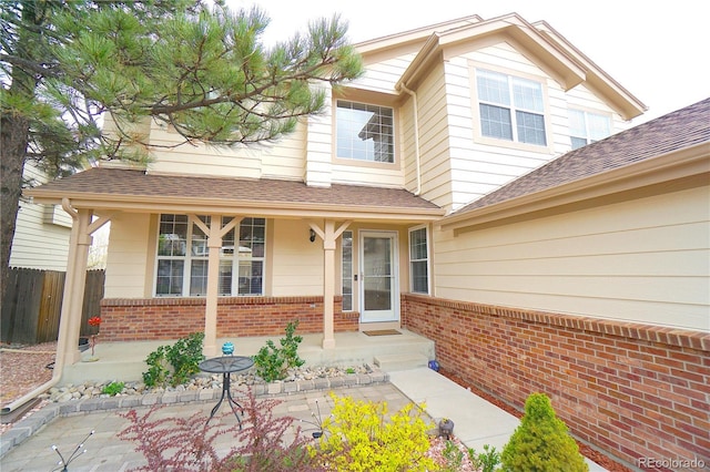entrance to property featuring a porch, fence, a shingled roof, and brick siding