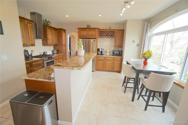 kitchen featuring wall chimney exhaust hood, stainless steel appliances, brown cabinets, and arched walkways