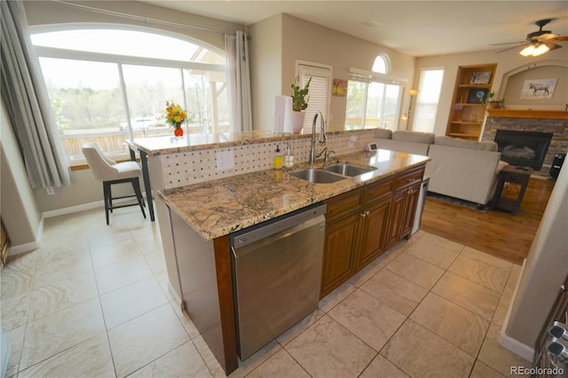 kitchen featuring a sink, brown cabinetry, a kitchen island with sink, stainless steel dishwasher, and light stone countertops