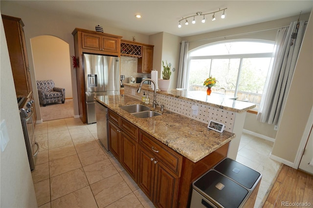kitchen featuring an island with sink, a sink, stainless steel appliances, brown cabinetry, and arched walkways