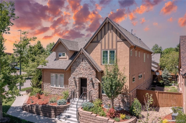 tudor home with stone siding, roof with shingles, fence, and stucco siding