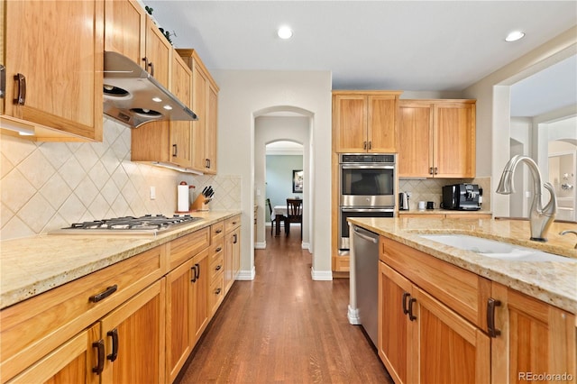 kitchen with arched walkways, light stone counters, under cabinet range hood, stainless steel appliances, and a sink