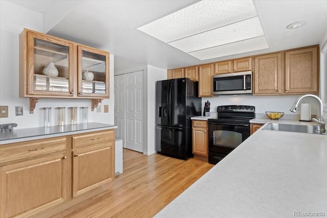 kitchen featuring glass insert cabinets, light wood-style flooring, black appliances, a sink, and light countertops