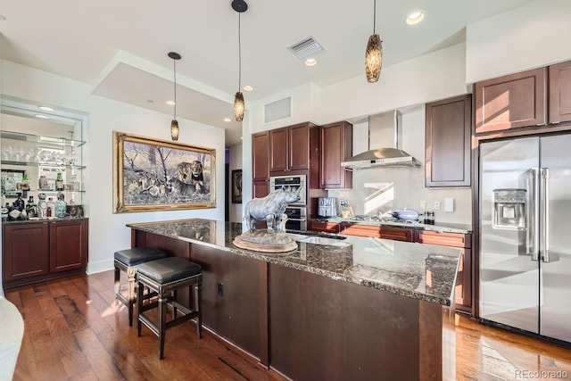 kitchen featuring wall chimney range hood, stainless steel appliances, dark hardwood / wood-style floors, decorative light fixtures, and dark stone counters