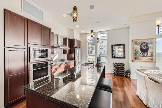 kitchen with pendant lighting, stainless steel appliances, dark wood-type flooring, a sink, and wall chimney exhaust hood