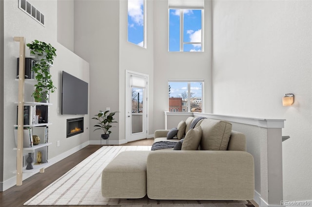 living area featuring baseboards, visible vents, a glass covered fireplace, a towering ceiling, and wood finished floors