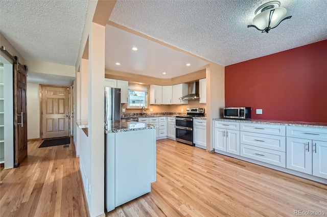 kitchen with dark stone counters, a barn door, appliances with stainless steel finishes, light wood-style floors, and wall chimney exhaust hood