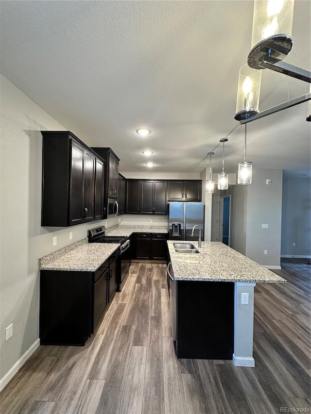 kitchen with hardwood / wood-style flooring, appliances with stainless steel finishes, hanging light fixtures, light stone counters, and an island with sink