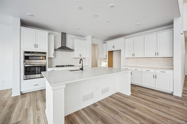 kitchen featuring stainless steel double oven, white cabinets, wall chimney exhaust hood, and a center island with sink