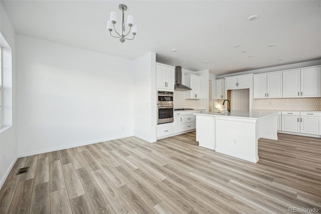 kitchen with sink, a kitchen island with sink, white cabinetry, decorative backsplash, and wall chimney exhaust hood