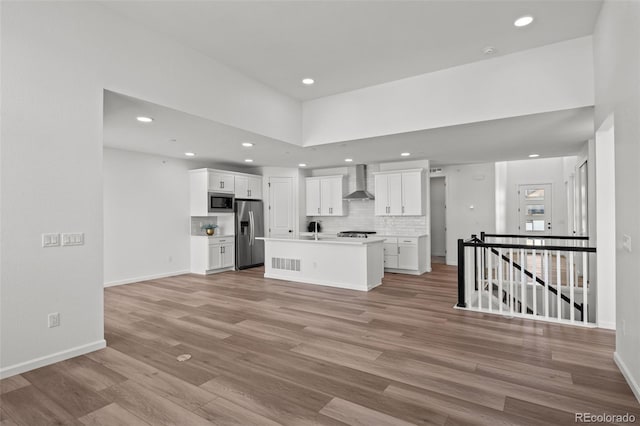 kitchen featuring light hardwood / wood-style flooring, appliances with stainless steel finishes, wall chimney range hood, a kitchen island with sink, and white cabinets