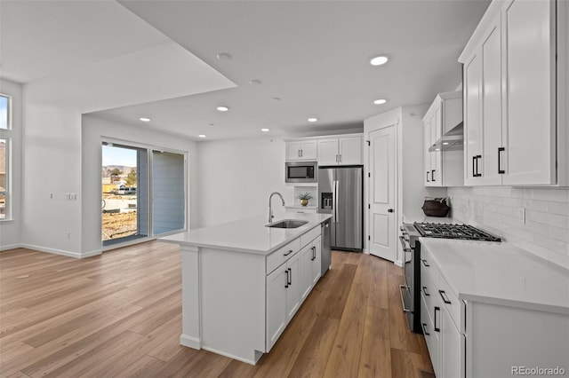 kitchen with sink, wall chimney range hood, stainless steel appliances, white cabinets, and a center island with sink