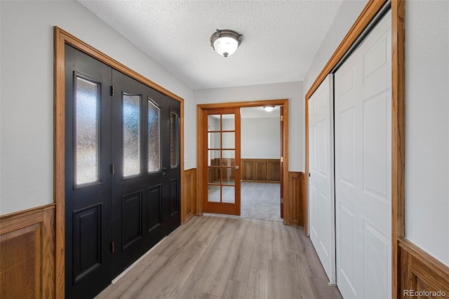 entrance foyer featuring french doors, a wainscoted wall, light wood-style flooring, a textured ceiling, and wooden walls