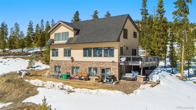 snow covered back of property with stone siding, stucco siding, a wooden deck, and roof with shingles