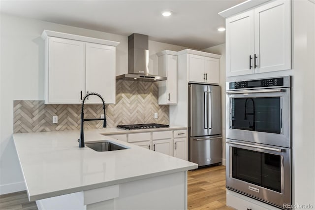 kitchen with stainless steel appliances, white cabinetry, a sink, a peninsula, and wall chimney exhaust hood