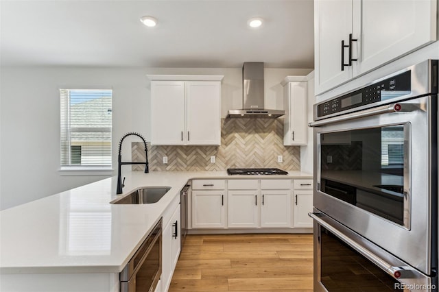 kitchen with stainless steel appliances, light countertops, white cabinetry, a sink, and wall chimney exhaust hood