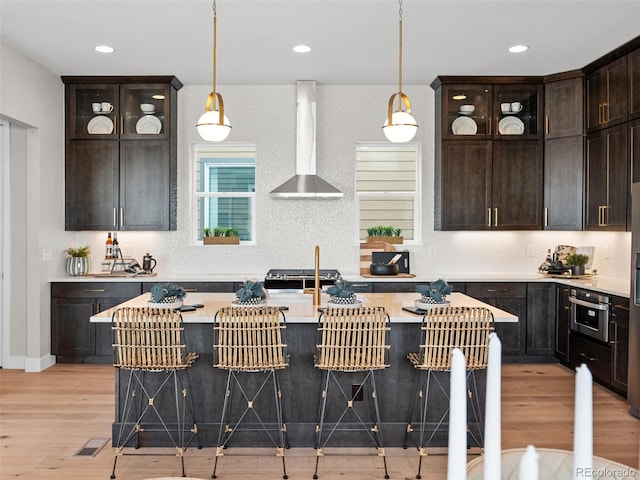 kitchen with stainless steel appliances, wall chimney range hood, a kitchen breakfast bar, light hardwood / wood-style floors, and a kitchen island with sink