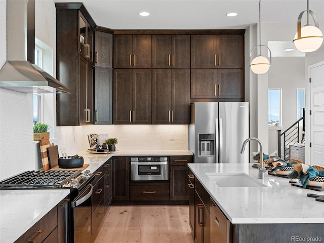 kitchen with wall chimney exhaust hood, stainless steel appliances, sink, pendant lighting, and light hardwood / wood-style floors