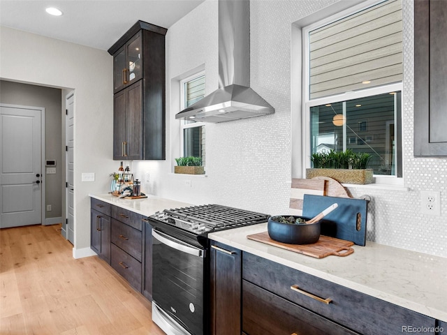 kitchen featuring light stone counters, stainless steel gas range oven, dark brown cabinetry, wall chimney range hood, and light hardwood / wood-style flooring