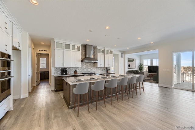 kitchen with white cabinets, light hardwood / wood-style floors, wall chimney exhaust hood, and an island with sink