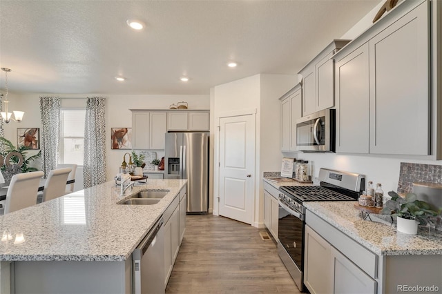 kitchen with light wood-type flooring, stainless steel appliances, a kitchen island with sink, sink, and hanging light fixtures