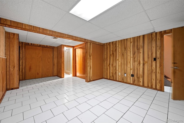 basement featuring light tile patterned floors, a drop ceiling, and wood walls