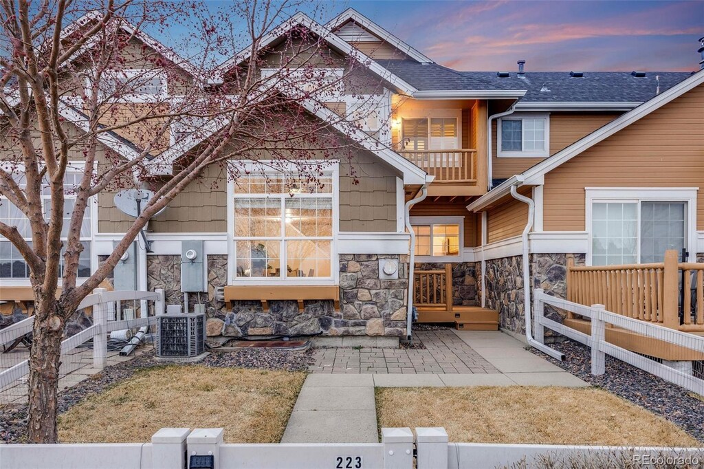 back of property at dusk featuring a balcony, cooling unit, fence, roof with shingles, and stone siding