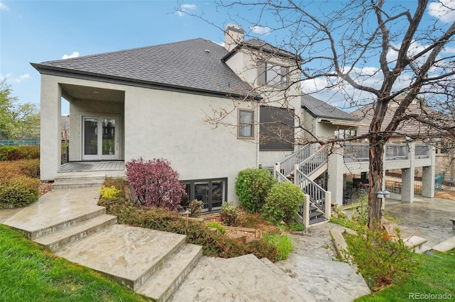 rear view of property with stairs, a shingled roof, stucco siding, a chimney, and french doors