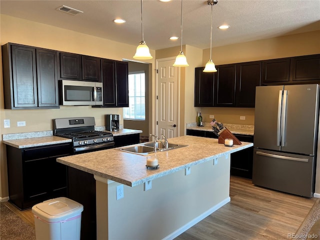 kitchen featuring pendant lighting, an island with sink, sink, stainless steel appliances, and light wood-type flooring