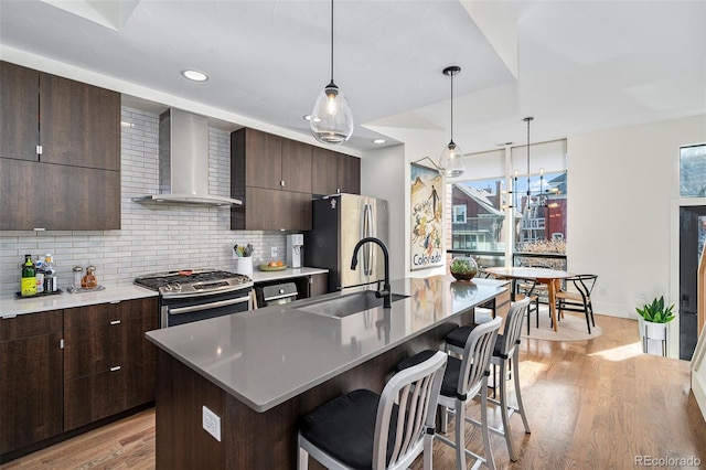 kitchen with wall chimney exhaust hood, sink, tasteful backsplash, stainless steel appliances, and a kitchen island with sink