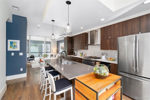 kitchen with wall chimney range hood, sink, a kitchen island with sink, stainless steel appliances, and decorative light fixtures