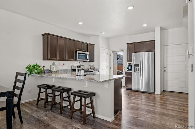 kitchen featuring stainless steel appliances, dark hardwood / wood-style floors, dark brown cabinetry, light stone countertops, and kitchen peninsula