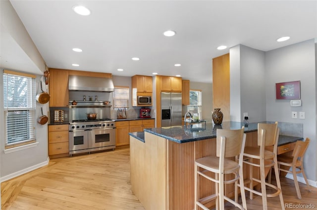 kitchen featuring a peninsula, stainless steel appliances, light wood-style floors, and wall chimney range hood