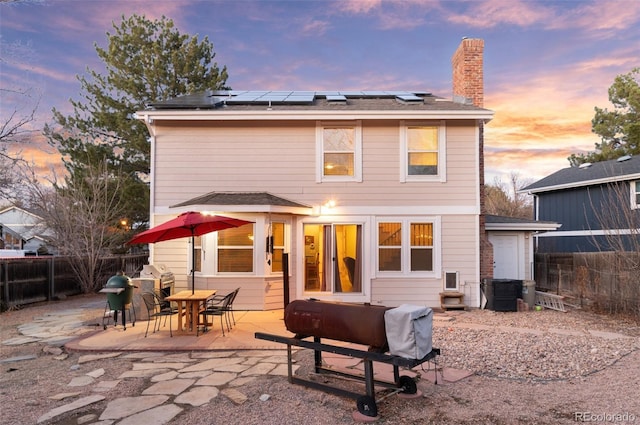 back of house at dusk featuring a patio area, fence, roof mounted solar panels, and a chimney