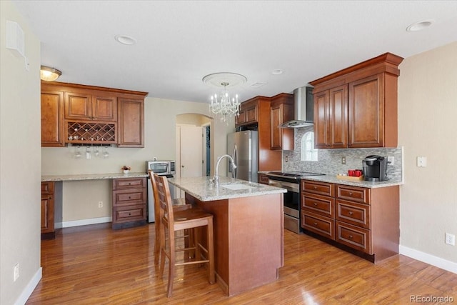 kitchen with light stone countertops, wall chimney exhaust hood, stainless steel appliances, sink, and a kitchen island with sink