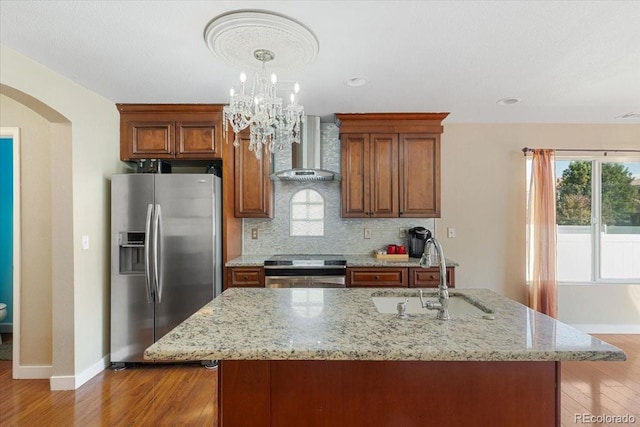 kitchen with light stone countertops, appliances with stainless steel finishes, wall chimney range hood, sink, and a center island with sink