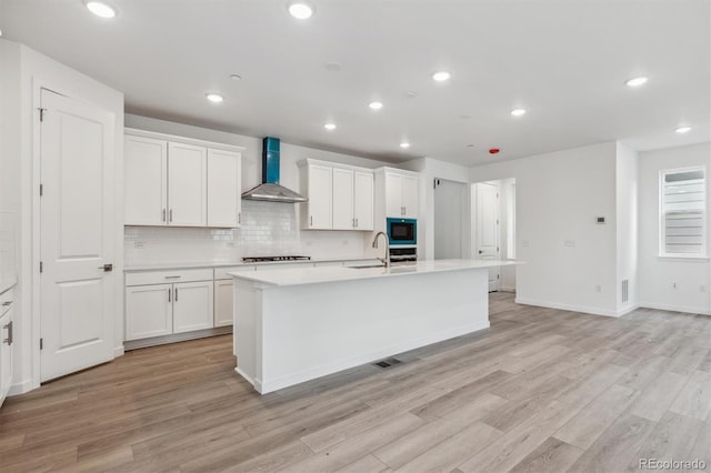 kitchen with wall chimney exhaust hood, black microwave, gas stovetop, a center island with sink, and light wood-style floors