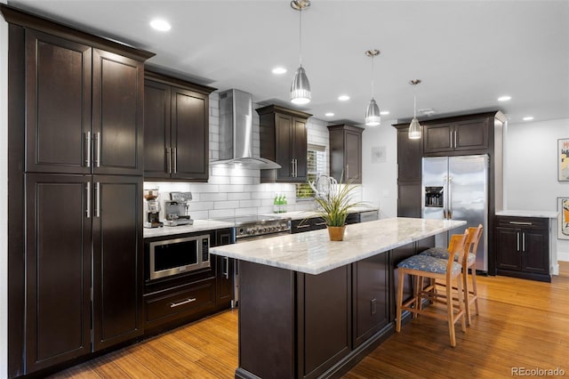 kitchen featuring appliances with stainless steel finishes, wall chimney exhaust hood, decorative backsplash, light wood-type flooring, and a kitchen island
