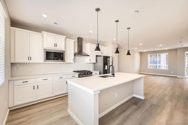 kitchen with appliances with stainless steel finishes, white cabinetry, sink, wall chimney range hood, and a center island with sink