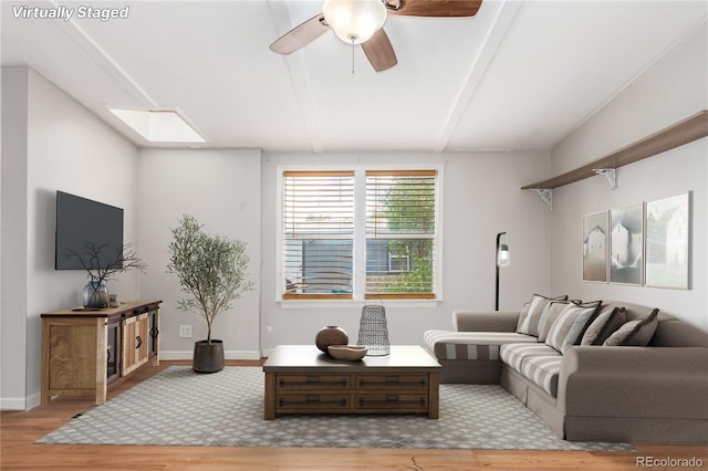 living room featuring ceiling fan, a skylight, wood finished floors, baseboards, and beamed ceiling