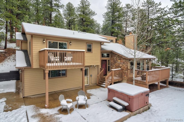 snow covered back of property with a hot tub, a chimney, a deck, a patio, and a balcony
