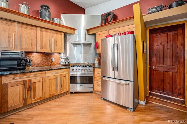 kitchen featuring stainless steel appliances, light hardwood / wood-style flooring, decorative backsplash, dark stone counters, and wall chimney range hood