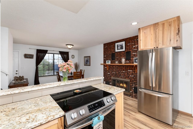 kitchen featuring a brick fireplace, light brown cabinetry, a textured ceiling, light wood-style flooring, and stainless steel appliances