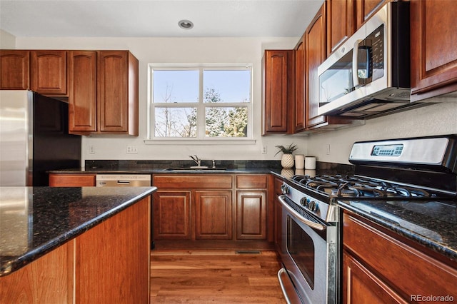 kitchen with appliances with stainless steel finishes, brown cabinetry, a sink, wood finished floors, and dark stone counters