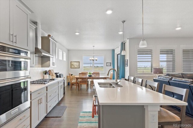 13476 Willow St, Thornton, CO 80602 - kitchen featuring stainless steel appliances, wall chimney range hood, a kitchen island with sink, light countertops, and dark wood finished floors