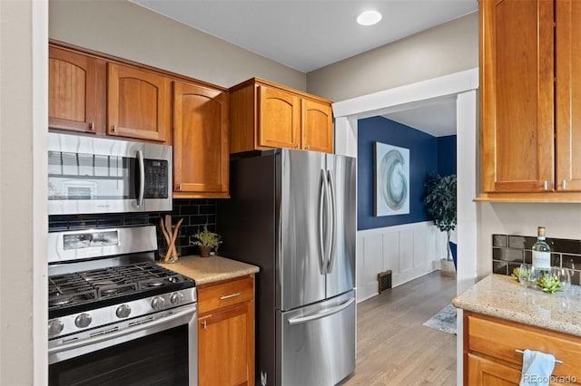 2271 Holly St, Denver, CO 80207 - kitchen featuring stainless steel appliances, light wood-style floors, a wainscoted wall, brown cabinets, and light stone countertops
