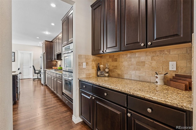 kitchen with dark brown cabinetry, tasteful backsplash, light stone counters, dark wood-style flooring, and stainless steel appliances